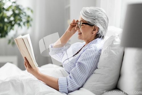 Image of old woman in glasses reading book in bed at home