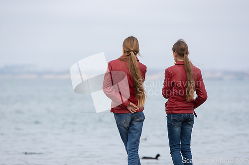 Image of Two girls in identical clothes look at the sea, close-up, view from the back