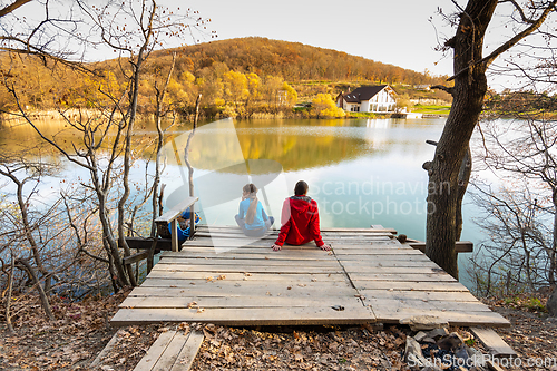 Image of A girl and a girl are sitting on a wooden bridge near a mountain lake, a view from the back of people