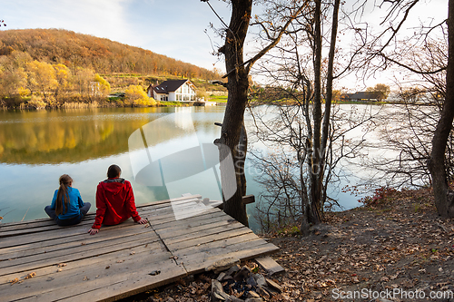 Image of A girl and a girl are sitting on a wooden bridge by the lake and looking at a beautiful autumn landscape