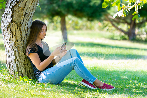 Image of A girl sits under a tree in a sunny park and looks at the smartphone screen