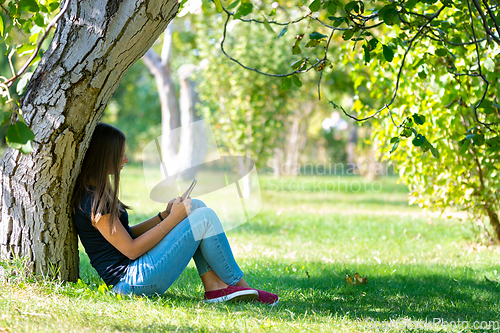 Image of A girl sits under a tree in a sunny park and looks at the phone screen