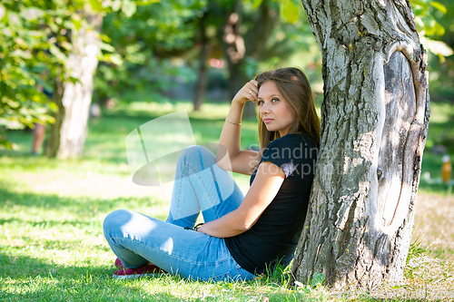 Image of A girl sits under a tree in a sunny park and looks into the distance in thought