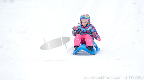 Image of Cheerful girl riding a sled downhill on a snow covered sledge trail in a white sunny winter mountain landscape