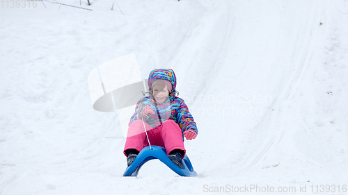 Image of Cheerful girl riding a sled downhill on a snow covered sledge trail in a white sunny winter mountain landscape