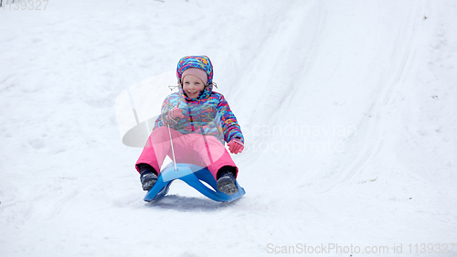 Image of Cheerful girl riding a sled downhill on a snow covered sledge trail in a white sunny winter mountain landscape