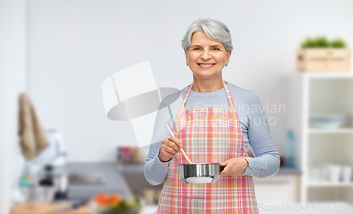 Image of old woman in kitchen apron with pot cooking food