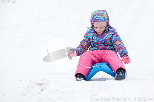 Image of Cheerful girl riding a sled downhill on a snow covered sledge trail in a white sunny winter mountain landscape