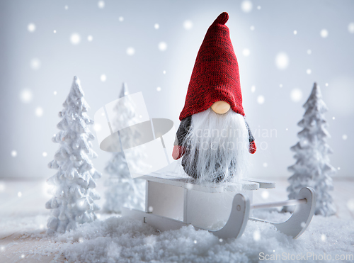 Image of Cute Christmas gnome is sitting on sled, winter country with white trees 