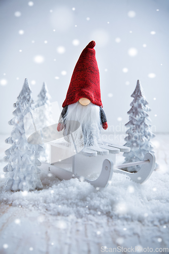 Image of Cute Christmas gnome is sitting on sled, winter country with white trees 