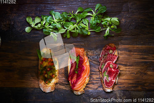 Image of Assorted bruschetta with roast beef, vegetables and lightly salted salmon with greens leaves on wooden background.