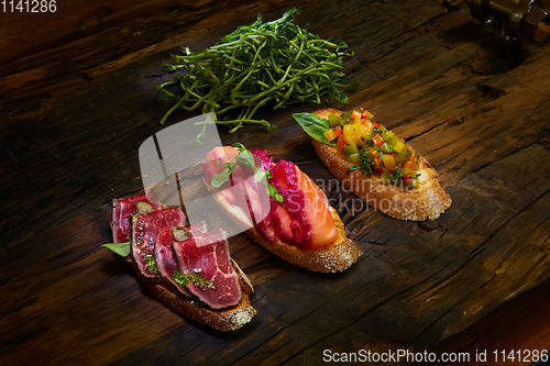 Image of Assorted bruschetta with roast beef, vegetables and lightly salted salmon with greens leaves on wooden background.