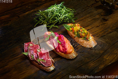 Image of Assorted bruschetta with roast beef, vegetables and lightly salted salmon with greens leaves on wooden background.