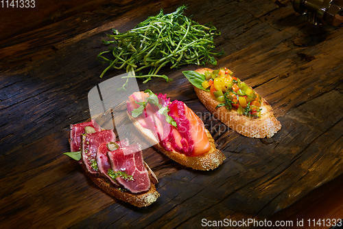 Image of Assorted bruschetta with roast beef, vegetables and lightly salted salmon with greens leaves on wooden background.