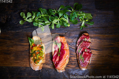 Image of Assorted bruschetta with roast beef, vegetables and lightly salted salmon with greens leaves on wooden background.