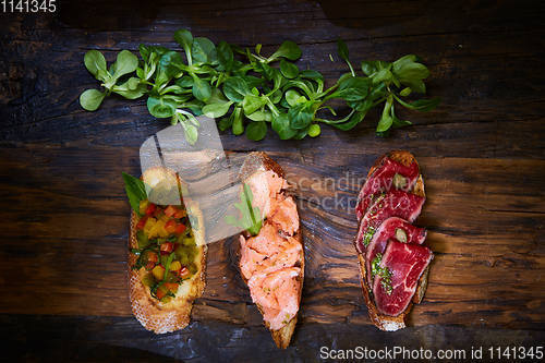 Image of Assorted bruschetta with roast beef, vegetables and lightly salted salmon with greens leaves on wooden background.