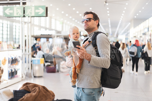 Image of Father traveling with child, holding his infant baby boy at airport terminal, checking flight schedule, waiting to board a plane. Travel with kids concept.
