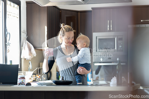 Image of Happy mother holding her little infant baby boy while drinking morning coffee and making pancakes for breakfast in domestic kitchen. Family lifestyle, domestic life concept.