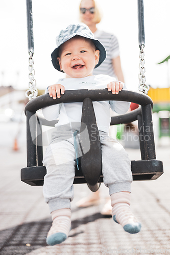 Image of Mother pushing her infant baby boy child on a swing on playground outdoors.