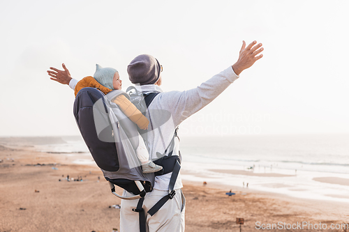 Image of Young father rising hands to the sky while enjoying pure nature carrying his infant baby boy son in backpack on windy sandy beach. Family travel concept.