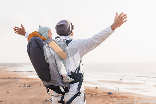 Image of Young father rising hands to the sky while enjoying pure nature carrying his infant baby boy son in backpack on windy sandy beach. Family travel concept.