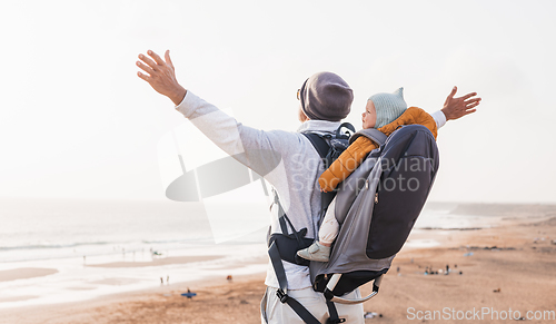 Image of Young father rising hands to the sky while enjoying pure nature carrying his infant baby boy son in backpack on windy sandy beach. Family travel concept.