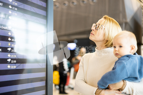 Image of Mother traveling with child, holding his infant baby boy at airport terminal, checking flight schedule, waiting to board a plane. Travel with kids concept.