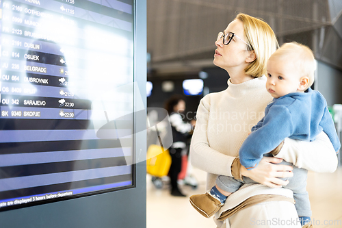 Image of Mother traveling with child, holding his infant baby boy at airport terminal, checking flight schedule, waiting to board a plane. Travel with kids concept.