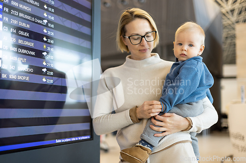 Image of Mother traveling with child, holding his infant baby boy at airport terminal, checking flight schedule, waiting to board a plane. Travel with kids concept.