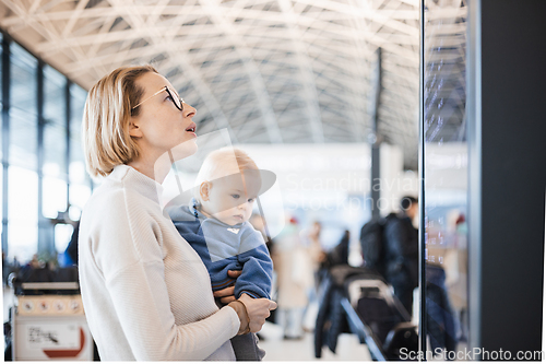 Image of Mother traveling with child, holding his infant baby boy at airport terminal, checking flight schedule, waiting to board a plane. Travel with kids concept.