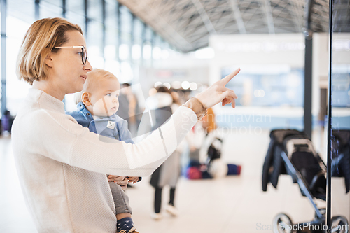 Image of Mother traveling with child, holding his infant baby boy at airport terminal, checking flight schedule, waiting to board a plane. Travel with kids concept.