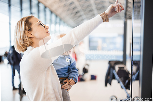 Image of Mother traveling with child, holding his infant baby boy at airport terminal, checking flight schedule, waiting to board a plane. Travel with kids concept.