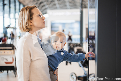 Image of Mother traveling with child, holding his infant baby boy at airport terminal, checking flight schedule, waiting to board a plane. Travel with kids concept.