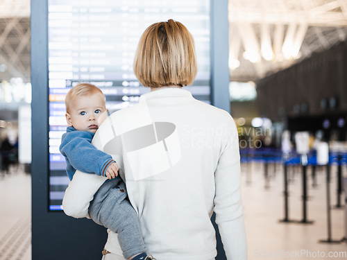 Image of Mother traveling with child, holding his infant baby boy at airport terminal, checking flight schedule, waiting to board a plane. Travel with kids concept.