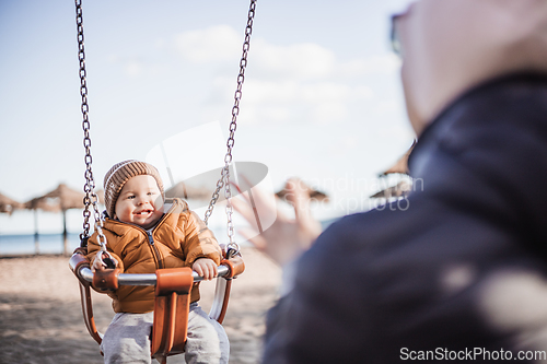 Image of Mother pushing her cheerful infant baby boy child on a swing on sandy beach playground outdoors on nice sunny cold winter day in Malaga, Spain.