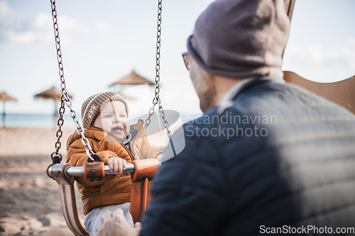 Image of Father pushing hir cheerful infant baby boy child on a swing on sandy beach playground outdoors on nice sunny cold winter day in Malaga, Spain.