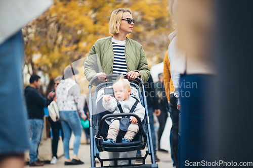 Image of Mother waling and pushing his infant baby boy child in stroller in crowd of people wisiting sunday flea market in Malaga, Spain.