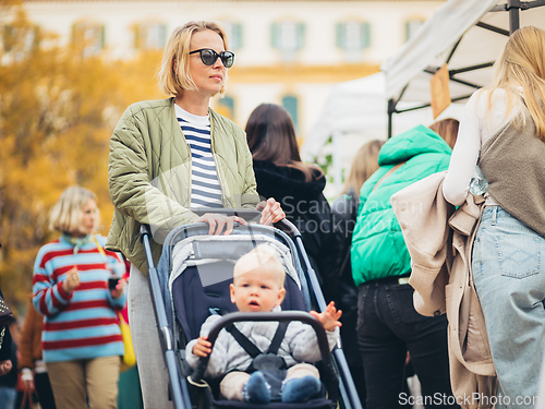 Image of Mother waling and pushing his infant baby boy child in stroller in crowd of people wisiting sunday flea market in Malaga, Spain.