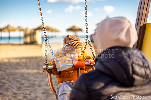 Image of Mother pushing her infant baby boy child on a swing on sandy beach playground outdoors on nice sunny cold winter day in Malaga, Spain.