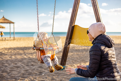 Image of Mother pushing her infant baby boy child on a swing on sandy beach playground outdoors on nice sunny cold winter day in Malaga, Spain.