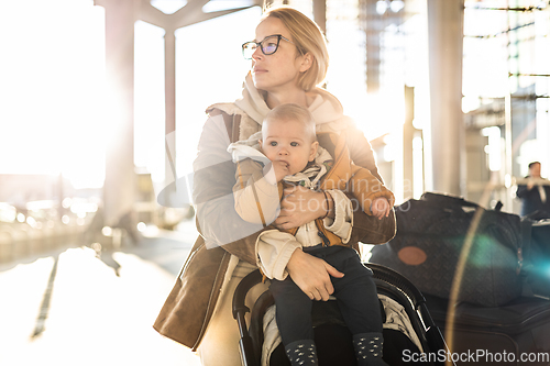 Image of Motherat travelling with his infant baby boy child, walking, pushing baby stroller and luggage cart in front of airport terminal station.