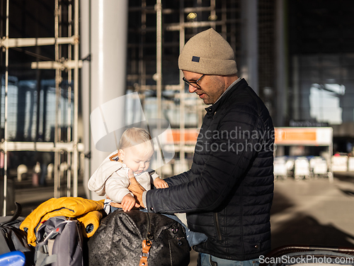 Image of Fatherat comforting his tired infant baby boy child sitting on top of luggage cart in front of airport terminal station while traveling wih family.