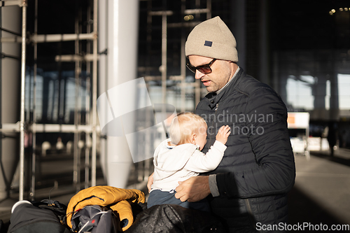 Image of Fatherat comforting his crying infant baby boy child tired sitting on top of luggage cart in front of airport terminal station while traveling wih family.