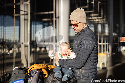 Image of Fatherat comforting his crying infant baby boy child tired sitting on top of luggage cart in front of airport terminal station while traveling wih family.