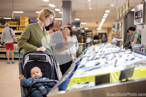 Image of Casualy dressed mother choosing fish in the fish market department of supermarket grocery store with her infant baby boy child in stroller.