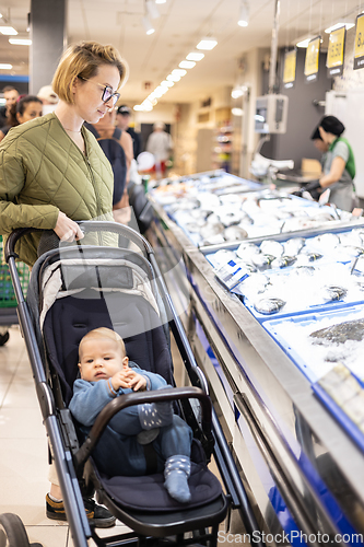 Image of Casualy dressed mother choosing fish in the fish market department of supermarket grocery store with her infant baby boy child in stroller.