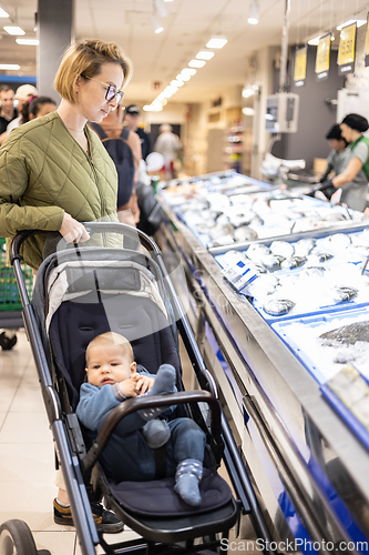 Image of Casualy dressed mother choosing fish in the fish market department of supermarket grocery store with her infant baby boy child in stroller.