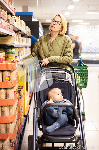 Image of Casualy dressed mother choosing canned products in department of supermarket grocery store with her infant baby boy child in stroller.