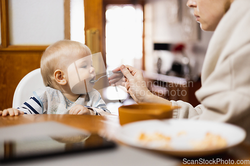 Image of Mother spoon feeding her infant baby boy child sitting in high chair at the dining table in kitchen at home