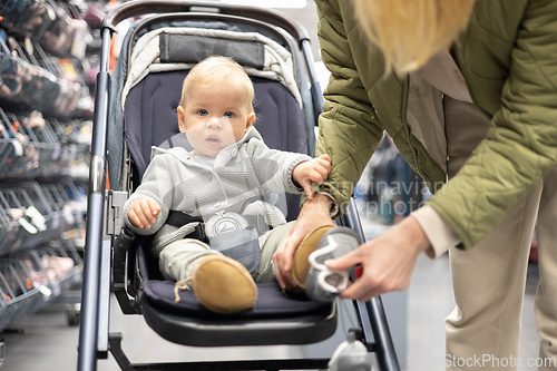 Image of Casualy dressed mother choosing sporty shoes and clothes products in sports department of supermarket store with her infant baby boy child in stroller.